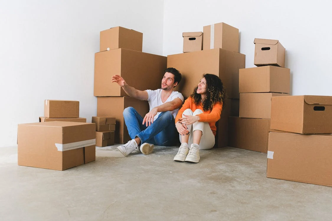 pexels-photo-7203842 A smiling couple sits on the floor, surrounded by stacked moving boxes. The scene suggest they are moving or right-sizing